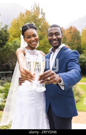 Portrait de mariée afro-américaine heureuse et marié grillant avec du champagne dans le jardin ensoleillé Banque D'Images