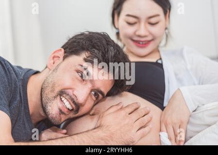 Père entendant son fils ou sa fille donner des coups de pied vérifier à l'intérieur du ventre de la mère lorsqu'il est assis sur le lit à la maison. Famille en bonne santé Banque D'Images