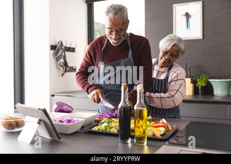 Heureux couple biracial senior portant des tabliers préparant des légumes dans la cuisine à la maison Banque D'Images