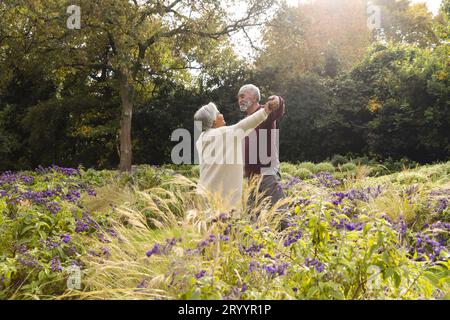 Heureux couple biracial senior dansant avec des fleurs autour d'eux dans le jardin à la maison Banque D'Images