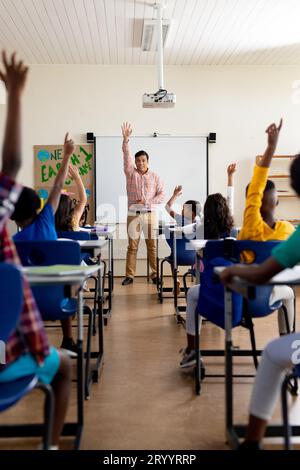 Enseignant masculin diversifié et écoliers du primaire levant la main en classe Banque D'Images