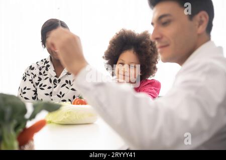 Médecin nutritionniste hispanique consultant un enfant métis avec sa mère afro-américaine au sujet d'une nutrition. Nourriture de légumes - une nouvelle façon pour le bien Banque D'Images