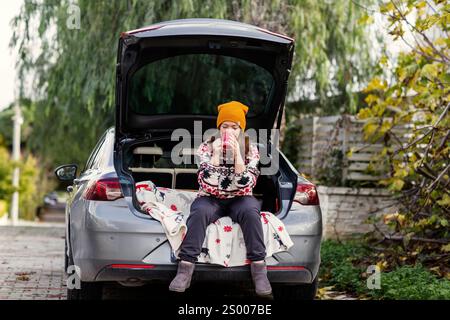 Femme en bonnet jaune assise avec du café dans le coffre de voiture Banque D'Images