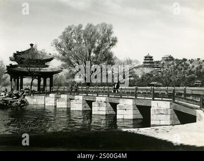 Beauté intemporelle du Palais d'été : pont panoramique traversant une eau tranquille avec une pagode traditionnelle et des arbres ornementaux dans un paysage chinois historique. Pékin, Chine, années 1930 Banque D'Images