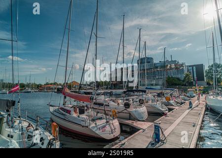 Une marina animée avec des voiliers amarrés sur des quais en bois, avec de hauts mâts, une eau calme et un bâtiment moderne en bord de mer sous un ciel bleu clair. Réf Banque D'Images