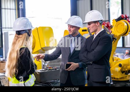 Trois professionnels s’engagent dans la discussion dans un cadre industriel rempli de machines. Deux hommes en costumes et une femme en équipement de sécurité partagent leurs idées, s. Banque D'Images