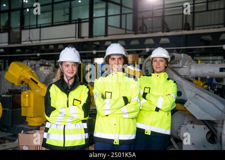 Trois femmes portent des vestes de sécurité brillantes et des casques de sécurité tout en se tenant debout avec les bras croisés à l'intérieur d'une usine de fabrication moderne. Banque D'Images