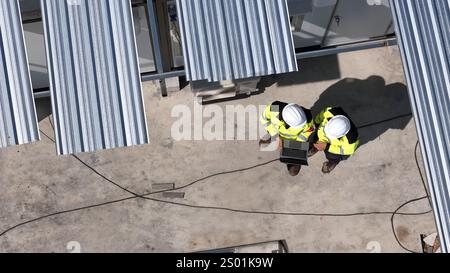 Deux ouvriers de la construction portant des casques de sécurité et des vestes réfléchissantes examinent les plans du projet sur un ordinateur portable. Ils sont situés sur un chantier de construction avec Banque D'Images