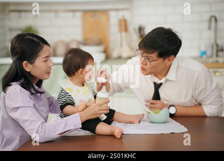 Une mère aide son partenaire à nourrir son bébé, créant un environnement chaleureux et nourrissant dans une cuisine bien éclairée pendant les repas. Banque D'Images