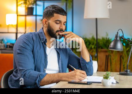 Indien confus homme freelance travaillant avec des documents du bureau à domicile, analysant l'information, lisant des documents d'affaires préparer le rapport financier. Jeune homme hispanique comptable analysant des documents graphiques Banque D'Images