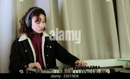 Une fille avec des écouteurs aime travailler sur un mixeur sonore, son expression concentrée et joyeuse. La photo met en lumière les thèmes de la production audio, de l'apprentissage Banque D'Images