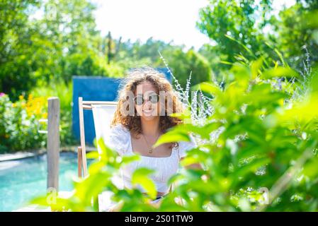 fröhliche Junge Frau mit Sonnenbrille sitzt im Liegestuhl am piscine *** jeune femme joyeuse avec des lunettes de soleil assise dans une chaise longue à la piscine Banque D'Images