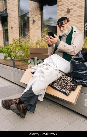 Portrait vertical d'un homme plus âgé assis sur le banc de la ville, en tapant smartphone au milieu de la solitude urbaine, éclairant la pauvreté et l'isolement dans le vieillissement Banque D'Images