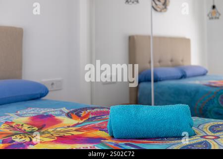 Cette chambre décorée de couleurs vives comprend une serviette roulée bleue sur un couvre-lit fleuri aux couleurs vives, une armoire à miroirs et une tête de lit beige. Banque D'Images