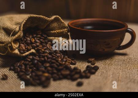 Une tasse de café noir en céramique brune placée à côté d'un sac en toile de jute renversant des grains de café torréfiés sur une surface texturée en toile de jute. Banque D'Images