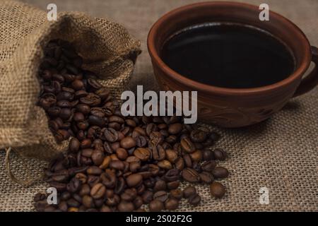 Une tasse en céramique brune rustique remplie de café noir placée à côté d'un sac en toile de jute renversant des grains de café torréfiés sur une surface en toile de jute. Banque D'Images