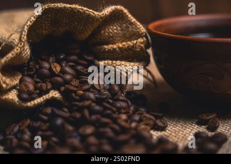 Vue rapprochée d'un sac en toile de jute renversant des grains de café torréfiés à côté d'une tasse de café noir en céramique brune sur une surface texturée en toile de jute. Banque D'Images