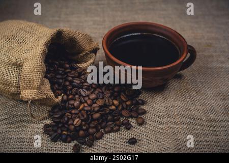 Une tasse en céramique brune rustique remplie de café noir à côté d'un sac en toile de jute renversant des grains de café torréfiés sur une surface en toile de jute. Banque D'Images