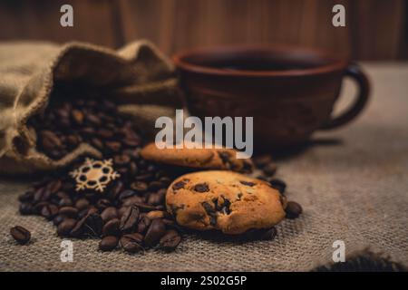 Gros plan de biscuits aux pépites de chocolat sur toile de jute avec des grains de café renversés d'un sac de toile de jute et une tasse de café noir en céramique brune rustique Banque D'Images