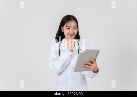 Une belle femme médecin asiatique positive dans une robe blanche se tient sur un fond blanc de studio, lisant des cas médicaux sur sa tablette numérique. Banque D'Images