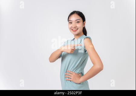 Une jeune femme asiatique souriante et insouciante pointant son bras avec un pansement adhésif en plâtre après avoir été vaccinée contre le virus, debout contre Banque D'Images
