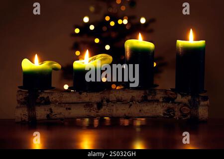 quatre bougies vertes allumées de l'avent devant un sapin de noël lumineux avec des lumières chaudes dans une pièce sombre Banque D'Images