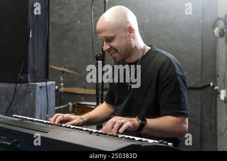 Producteur de son travaillant sur un nouveau hit dans un studio d'enregistrement, jouant au clavier avec une expression ciblée. Environnement professionnel avec batterie, microphone et équipement sonore. Banque D'Images