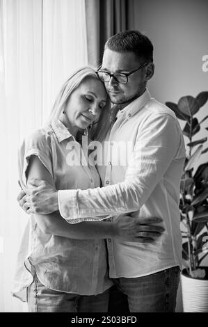 Couple heureux embrassant sur fond clair à la maison. Jolie femme souriant et embrassant son mari les yeux fermés. Couple doux en vêtements décontractés. Image en noir et blanc. Banque D'Images