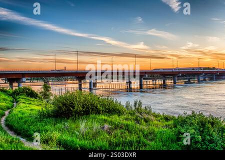 Un pont enjambe une rivière avec un beau coucher de soleil en arrière-plan. Le pont est entouré d'herbe verte luxuriante et d'arbres, créant un calme et serein Banque D'Images