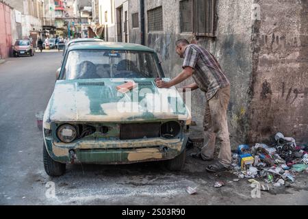 Un réparateur automobile sable une vieille voiture dans la rue d'Alexandrie, en Égypte, la préparant pour la peinture. Savoir-faire automobile urbain. Banque D'Images