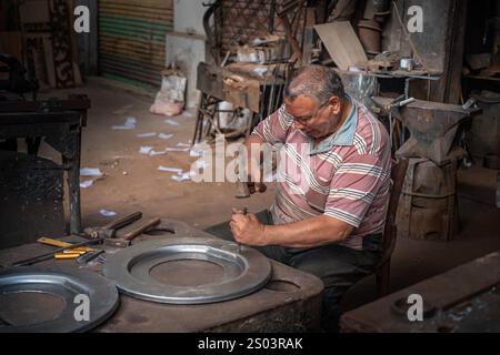 Un artisan à Alexandrie, en Egypte, travaillant dans un atelier, façonnant méticuleusement une plaque de métal à la main, entouré d'outils et de matériaux traditionnels. Banque D'Images