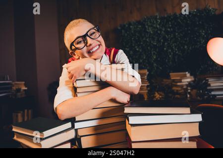 Jeune femme aux cheveux blonds courts, portant des lunettes et un gilet rouge, sourit en serrant une pile de livres. Banque D'Images