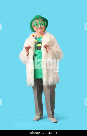Femme âgée heureuse en manteau de fourrure, lunettes et chapeau leprechaun pour la célébration de Patrick's Day sur fond bleu Banque D'Images