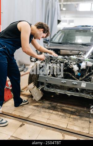 Vue arrière Vertica d'un mécanicien masculin concentré en uniforme bleu inspectant le moteur de la voiture dans l'atelier, soulignant la complexité du travail. Banque D'Images
