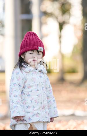 Une jeune fille japonaise de 3 ans portant un chapeau tricoté rouge et une veste en duvet blanche se tient avec des feuilles tombées par un après-midi froid d'hiver dans un étang de parc à Ha Banque D'Images