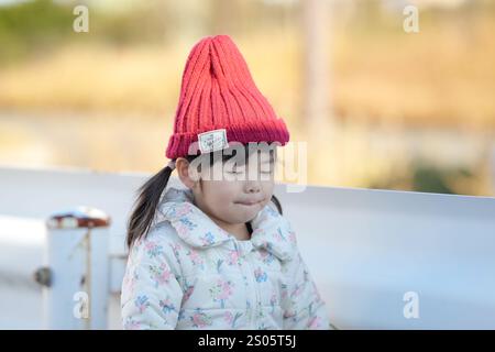 Une jeune fille japonaise de 3 ans portant un chapeau tricoté rouge et une veste en duvet blanche se tient à côté d'un garde-corps dans un parc de la préfecture de Fukuoka dans le wi froid Banque D'Images