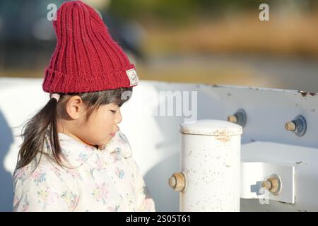 Une jeune fille japonaise de 3 ans portant un chapeau tricoté rouge et une veste en duvet blanche se tient à côté d'un garde-corps dans un parc de la préfecture de Fukuoka dans le wi froid Banque D'Images