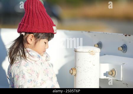 Une jeune fille japonaise de 3 ans portant un chapeau tricoté rouge et une veste en duvet blanche se tient à côté d'un garde-corps dans un parc de la préfecture de Fukuoka dans le wi froid Banque D'Images