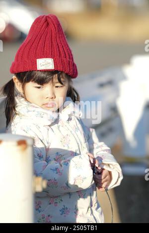 Une jeune fille japonaise de 3 ans portant un chapeau tricoté rouge et une veste en duvet blanche se tient à côté d'un garde-corps dans un parc de la préfecture de Fukuoka dans le wi froid Banque D'Images