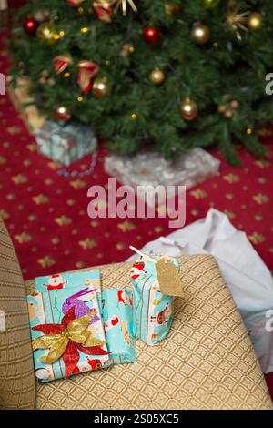 Cadeaux festifs avec un emballage coloré et des noeuds placés sur une chaise à motifs près d'un sapin de Noël décoré, avec des ornements et un décor de tapis rouge Banque D'Images