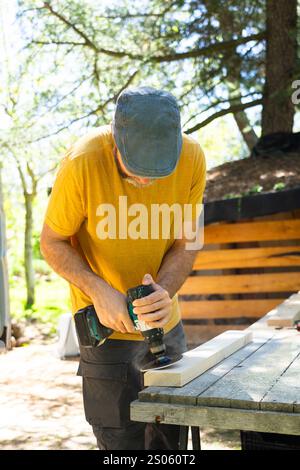 Carpenter utilise une ponceuse sans fil pour lisser la planche de bois à l'extérieur, avec la sciure de bois volante Banque D'Images