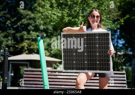 Femme souriante utilisant le panneau solaire portable pour charger son scooter. Jeune femme montrant le pouce jusqu'à la caméra. Femme heureuse de son style de vie vert. Banque D'Images