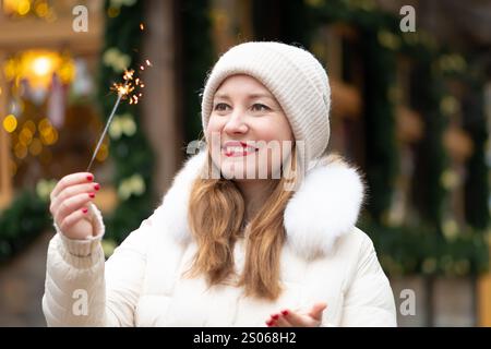 Une femme joyeuse tient un feu de Bengale étincelant et sourit joyeusement dehors pendant les vacances d'hiver entouré de décorations festives et de lumières chaudes Banque D'Images