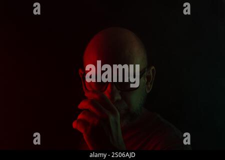 Portrait en gros plan de l'homme chauve et barbu dans des lunettes de prescription posant pour la photo. Isolé sur fond fumé foncé avec des lumières colorées. Ameri du Sud Banque D'Images