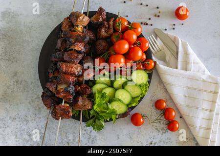 Vue de dessus de brochettes de viande grillée avec tomates cerises fraîches, tranches de concombre et persil sur une assiette noire, accompagnées de couverts enveloppés dans une serviette Banque D'Images