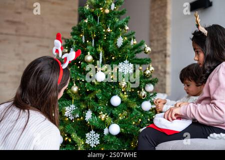 Une femme et deux enfants décorent un arbre de Noël. La femme porte un bandeau de renne et les enfants Banque D'Images