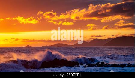 Un magnifique coucher de soleil sur l'océan, avec des teintes orange et jaune éclatantes illuminant le ciel. Les vagues s'écrasent contre les rivages rocheux, tandis que les îles lointaines Ar Banque D'Images