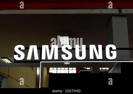 Signe lumineux blanc du logo de la marque Samsung brillant sur un fond sombre dans un magasin intérieur ou un cadre d'exposition. Banque D'Images