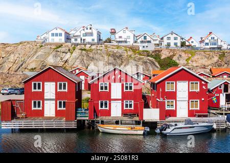 Trois maisons en bois peintes en rouge alignées le long du canal de la mer à Smögen, en Suède, avec des maisons blanches au sommet de la falaise derrière par une journée d'été ensoleillée. Banque D'Images