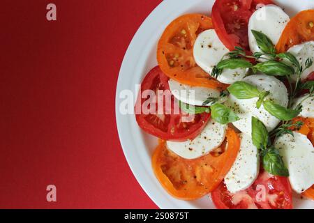 Salade caprese fraîche avec tomates tranchées, fromage mozzarella et feuilles de basilic, assaisonnée de poivre, servie sur une assiette blanche avec un bac rouge vif Banque D'Images